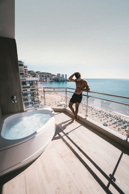 Full body of unrecognizable young shirtless male traveler in shorts leaning on railing and touching head while relaxing on terrace with picturesque sea view during summer holidays